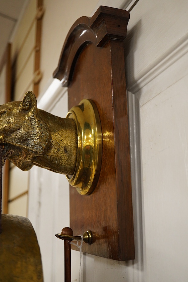 An early 20th century brass wall hanging gong, the holder in the form of a big cat, mounted on an oak plinth, the gong is supported on a bone clenched between its teeth, with the beater on a separate hook behind, 55cm to